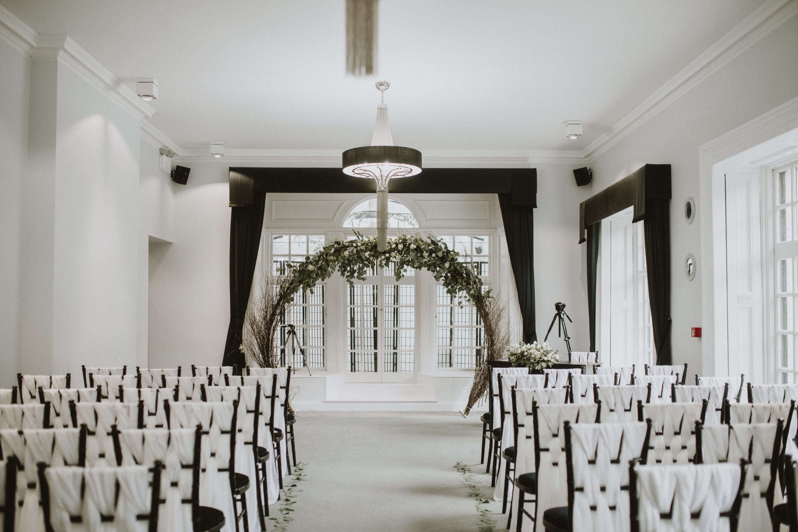 Wedding ceremony set in The Study at Swynford Manor with large floral arch in front of black chiavari chairs dressed with white tulle