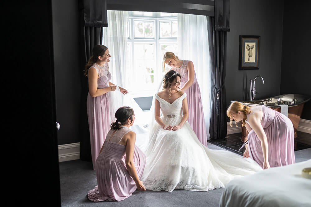 Bride in white wedding dress being attended by her four bridesmaids in pale pink in the bridal suite at Swynford Manor