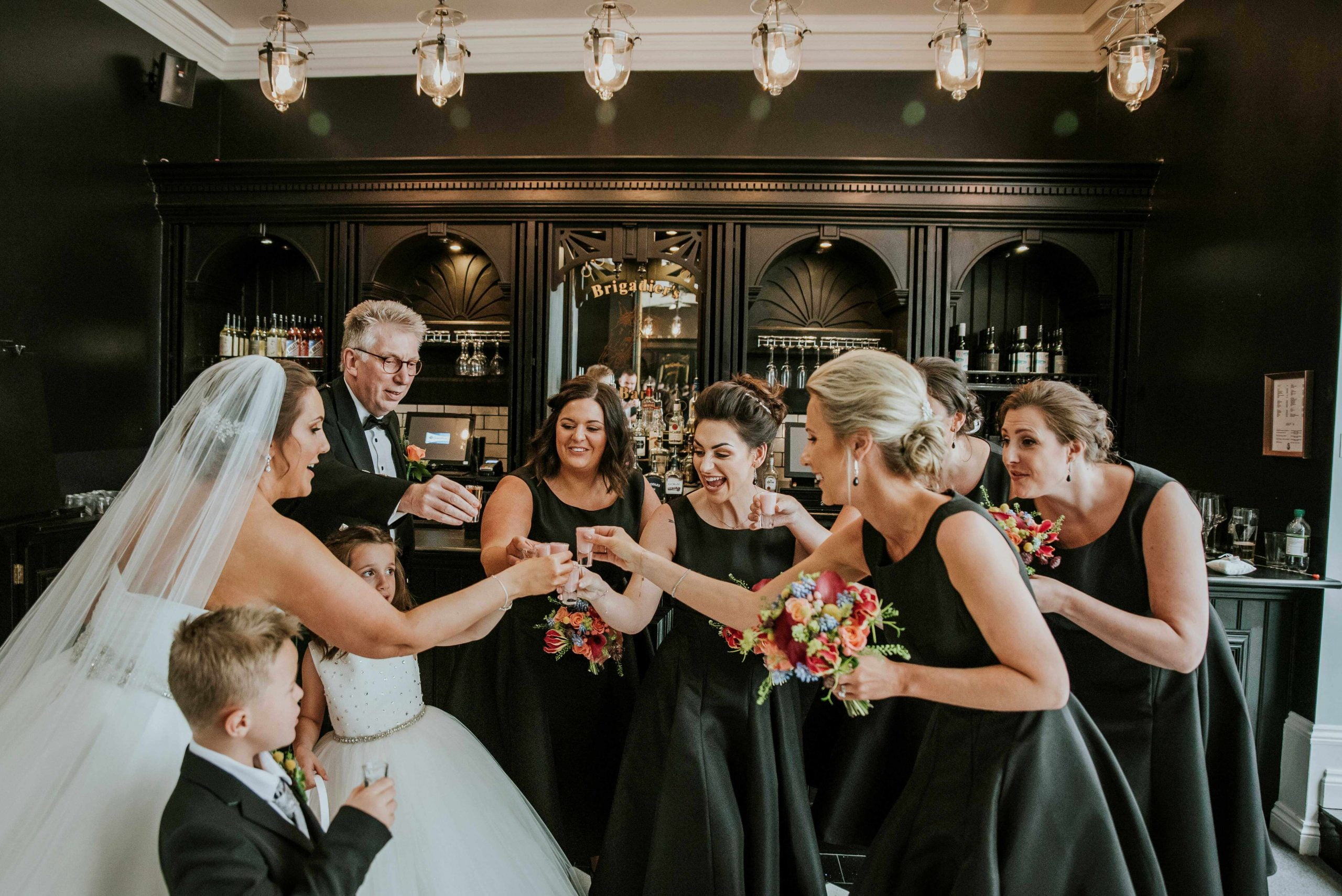 Bride cliking glasses with her bridesmaids in black dresses in the bar at Swynford Manor