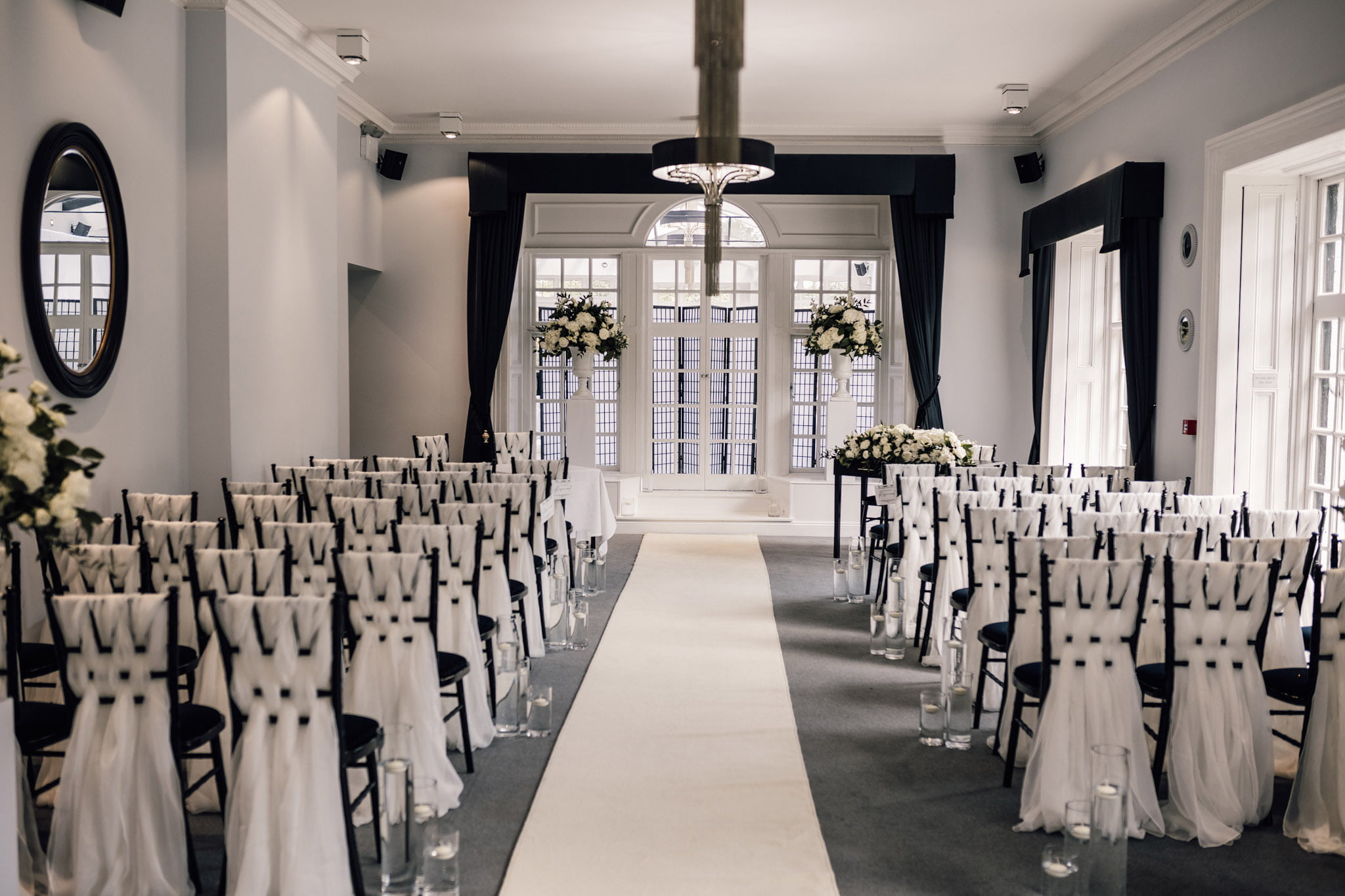 Ceremony setup in the Study at Swynford Manor with black chiavari chairs dressed with white voile facing french windows framed by black curtains and large white floral decorations