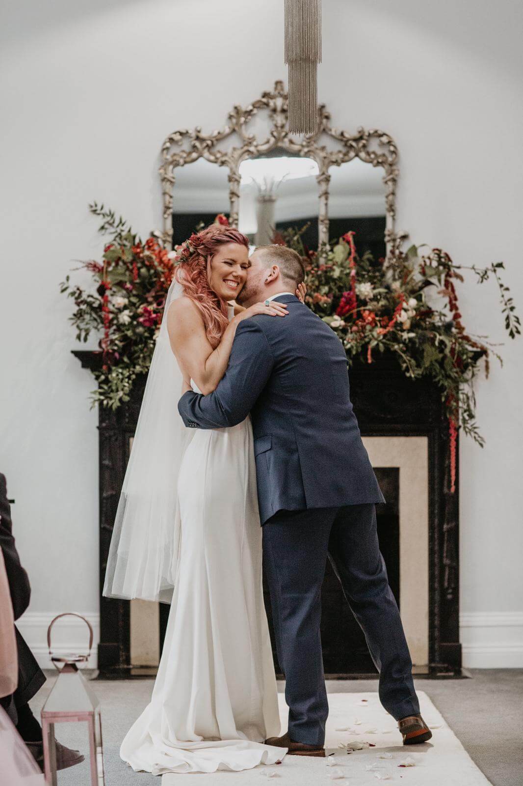 Bride and groom kissing after just getting married in front of the ornate fireplace with autumnal coloured flowers on the mantlepiece in the Study at Swynford Manor