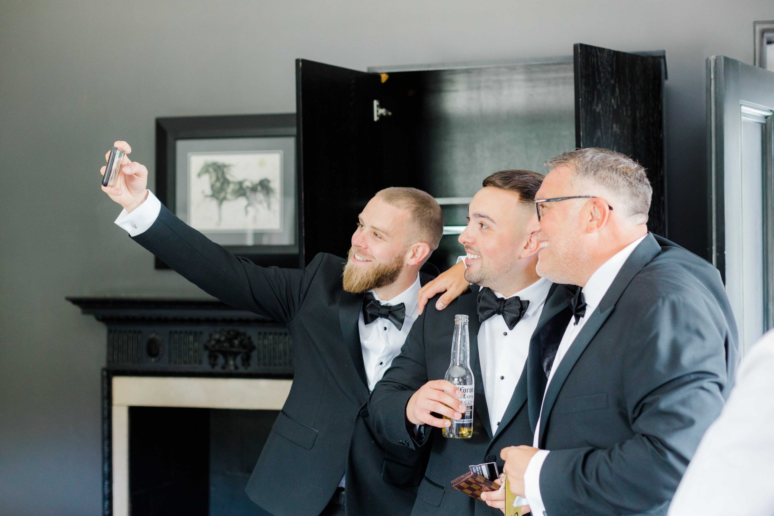 Groom, best man and his father having a selfie in front of the fireplace in a bedroom at Swynford Manor
